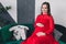 Young pregnant woman in red dress sitting in sofa in studio baby clothing.