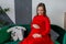 Young pregnant woman in red dress sitting in sofa in studio baby clothing. Belly; care