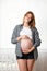 A young pregnant woman shows off her tummy and inflates a bubble of gum. White window in the background. Conception of