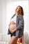 A young pregnant woman shows off her tummy and inflates a bubble of gum. White window in the background. Conception of