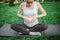 Young pregnant woman sit in lotus pose on yoga mate in park. She look at belly and hold hands there in heart shape.