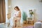 young pregnant woman is sitting in a chair in a bright room of her house and stroking her tummy