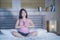 Young pretty Asian Chinese woman 20s or 30s relaxing at home bedroom sitting on bed in namaste yoga position in meditation and rel