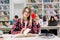 Young pretty blond woman reading book and drinking coffee at the table in modern college library. Multiracial students