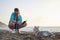 Young pretty blond woman, taking picture on her phone of her small husky puppy. Owner and the light grey dog, playing on the sandy