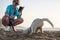 Young pretty blond woman, taking picture on her phone of her small husky puppy. Owner and the light grey dog, playing on the sandy