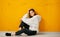 Young pretty calm relaxing girl in a white hoodie poses sitting on the floor against a yellow concrete wall.