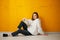 Young pretty calm relaxing girl in a white hoodie poses sitting on the floor against a yellow concrete wall.