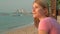 Young pretty dreamy woman in pink t-shirt on beach sea sunset boats cityscape at background