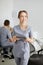 A young pretty female dentist stands in the middle of the office, and in the background a colleague is advising patients