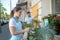 Young pretty female spraying plants outside flower shop, bearded female standing behind