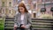 Young pretty ginger woman in jacket sitting on bench and using tablet, city street background