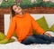 A young pretty girl is sitting on a bed in her room in a bright orange hooded sweatshirt or hoody