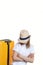 Young pretty girl tourist hiding face under stylish hat stands with hands crossed on chest near suitcase on wheels. Half-length