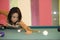 Young pretty and happy Asian girl playing snooker holding stick at pool table in night club or bar