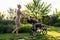 Young pretty mother with baby in stroller enjoying walking in green fresh garden at sunset. Mom having fun with baby in pram in be