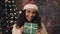 Young pretty mulatto woman at santas hat and decorative lights on her neck holding present box in her hands. She