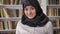 Young pretty muslim women in hijab looking into camera and smiling, standing in library, shelves with books background