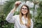 Young pretty smiling blond woman scientist agronomist wearing white lab coat, posing to camera in greenhouse with tropic