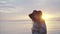 Young pretty and smiling woman in black hat and glasses near the sea at sunset