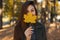 Young pretty stylish girl with blue eyes in a fashionable green coat, holding a yellow autumn leaf near the face while walking