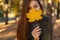 Young pretty stylish woman with blue eyes in a fashionable green coat, holding a yellow autumn leaf near the face