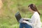 Young pretty successful smart business woman or student in light casual clothes sitting on grass using laptop in field