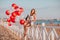 Young pretty woman holding champagne and glass sitting on a fence at the beach against balloons and sea