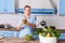 Young pretty woman holding jar of pickles and smiling looking at camera, cooking green salad in modern kitchen