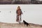 Young pretty woman with long wavy fair hair standing on sandy beach at river, playing with black dog Zwergpinscher.