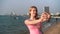 Young pretty woman in pink t-shirt taking selfie on beach sea sunset boats cityscape at background