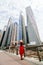 Young pretty woman in red dress and straw hat pointed with hands on downtown skycrapers in modern city. Low angle view