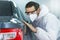 Young professional car painter wearing protective suit standing next to car in car painting room inspecting car body to look for