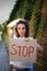 Young protesting woman in white shirt and jeans holds protest sign broadsheet placard with slogan `Stop` for public