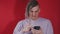 Young puzzled man browsing mobile phone on red background. Shocked guy with dreadlocks grimacing looking into smartphone