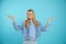 Young puzzled trendy blonde woman, teenager in eyeglasses and jeans denim jacket with outstretched arms on blue backview