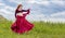 Young red-haired girl in red dance dress dances on green meadow against blue summer sky.