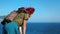 A young red-haired girl traveler with a cowboy hat and backpack stands on top of a mountain and looks down at the sea.