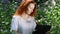 Young red-haired woman work on laptop computer among flowers in a city park