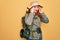 Young redhead backpacker woman hiking wearing backpack and hat over yellow background with hand on headache because stress