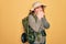 Young redhead backpacker woman hiking wearing backpack and hat over yellow background laughing and embarrassed giggle covering