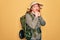 Young redhead backpacker woman hiking wearing backpack and hat over yellow background laughing nervous and excited with hands on