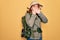 Young redhead backpacker woman hiking wearing backpack and hat over yellow background shocked covering mouth with hands for
