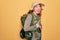 Young redhead backpacker woman hiking wearing backpack and hat over yellow background smiling and laughing hard out loud because