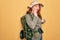 Young redhead backpacker woman hiking wearing backpack and hat over yellow background touching mouth with hand with painful