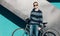 Young redhead man standing next to a vintage bicycle in sunglasses, sweater, jeance near turquoise wall warm summer sunny day