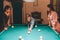 Young redhead player stand between two models. He aims into billiard ball. Young women stand and look at gamel. They lean to tabl