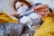 A young redhead woman looks at a thermometer next to a cat. Sick girl lies at home in a medical mask