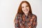 Young redhead woman with pale skin and white smile looking happy at camera, standing over white background