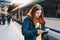 Young redhhead woman with backpack checking her ticket and using smartphone on background train. Railroad transport